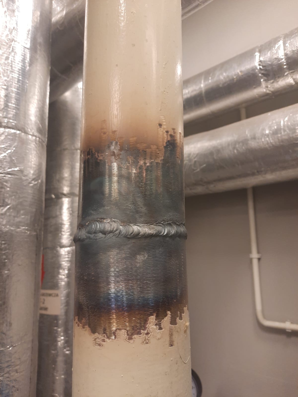 Spoina na pionowej rurze w pomieszczeniu technicznym. Widoczne ślady po spawaniu i czyszczeniu powierzchni. Inne rury w tle.