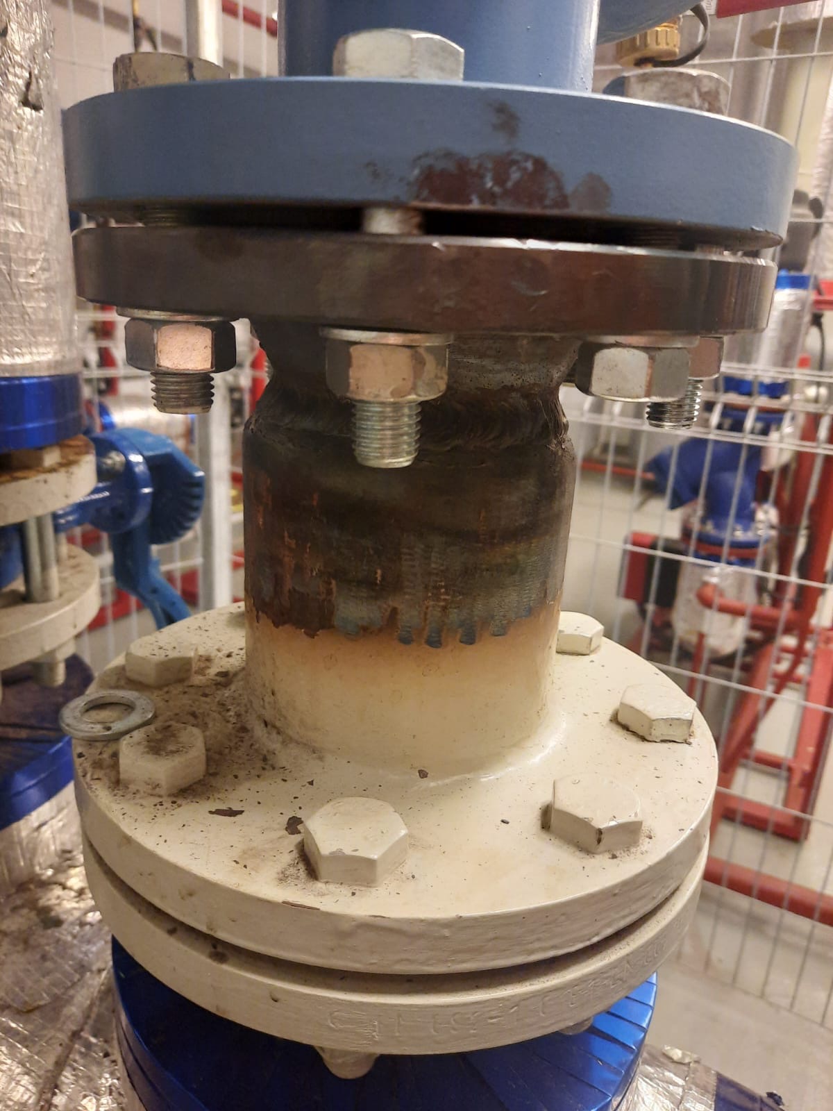 Szczegółowe ujęcie złącza kołnierzowego rury, z widocznymi śrubami i spoinami, sugerujące instalację hydrauliczną lub grzewczą w pomieszczeniu technicznym.