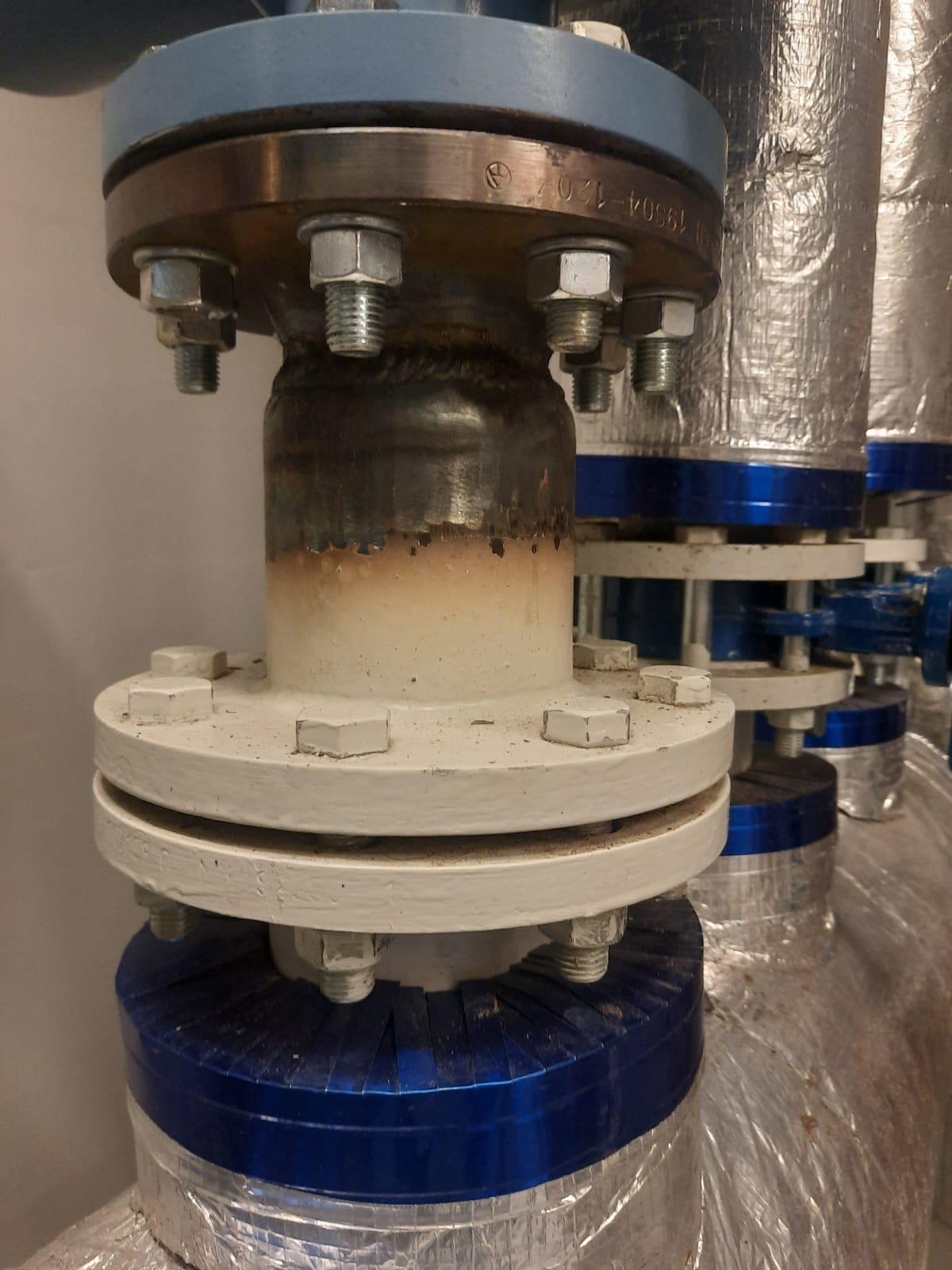 Spojenie rur z kołnierzami, śrubami i spawem, z izolacją termiczną na rurach w tle. Widoczne ślady spawania na rurze.