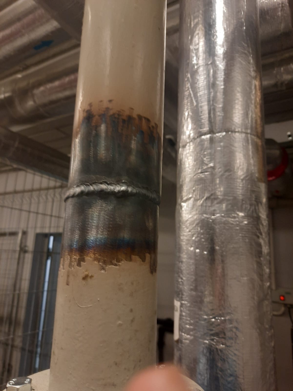Spoina na rurze instalacji centralnego ogrzewania, z widocznymi śladami spawania i przegrzania, obok rury izolowanej folią aluminiową. Detal z wnętrza kotłowni.