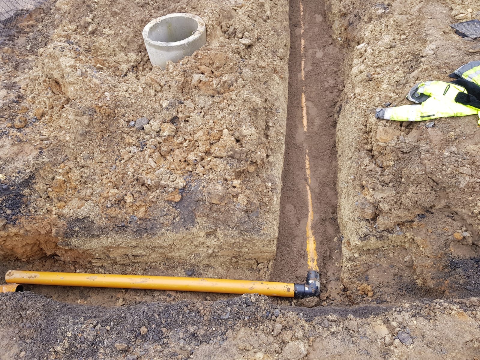 Żółta rura w wykopie, widoczny fragment studzienki betonowej i odzież robocza. Ujęcie z góry, prezentujące instalację podziemną w trakcie realizacji.