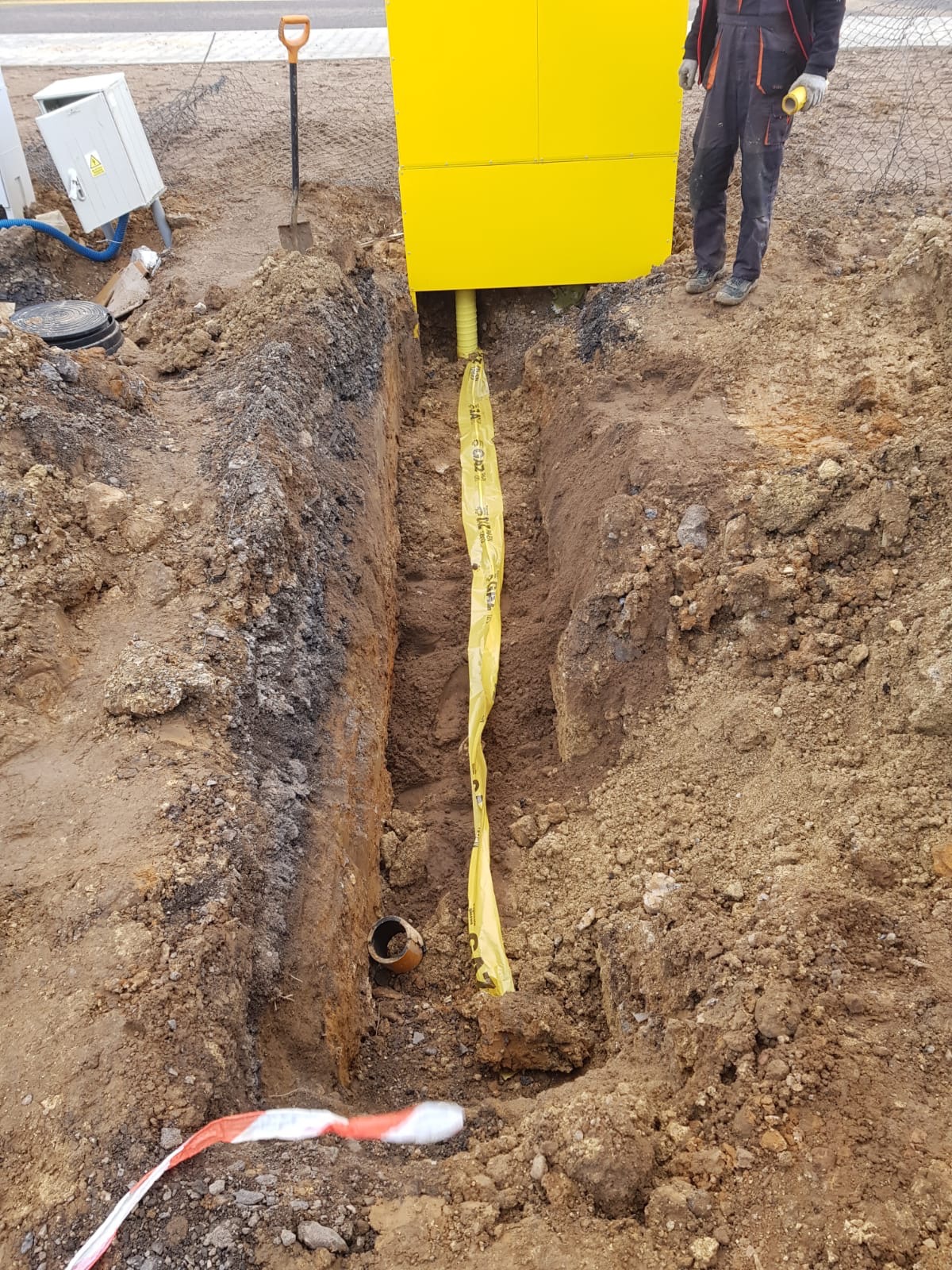 Wykop z żółtą rurą osłonową i taśmą ostrzegawczą w ziemi, obok żółta skrzynka i narzędzia. Widoczny fragment pracownika w kombinezonie.