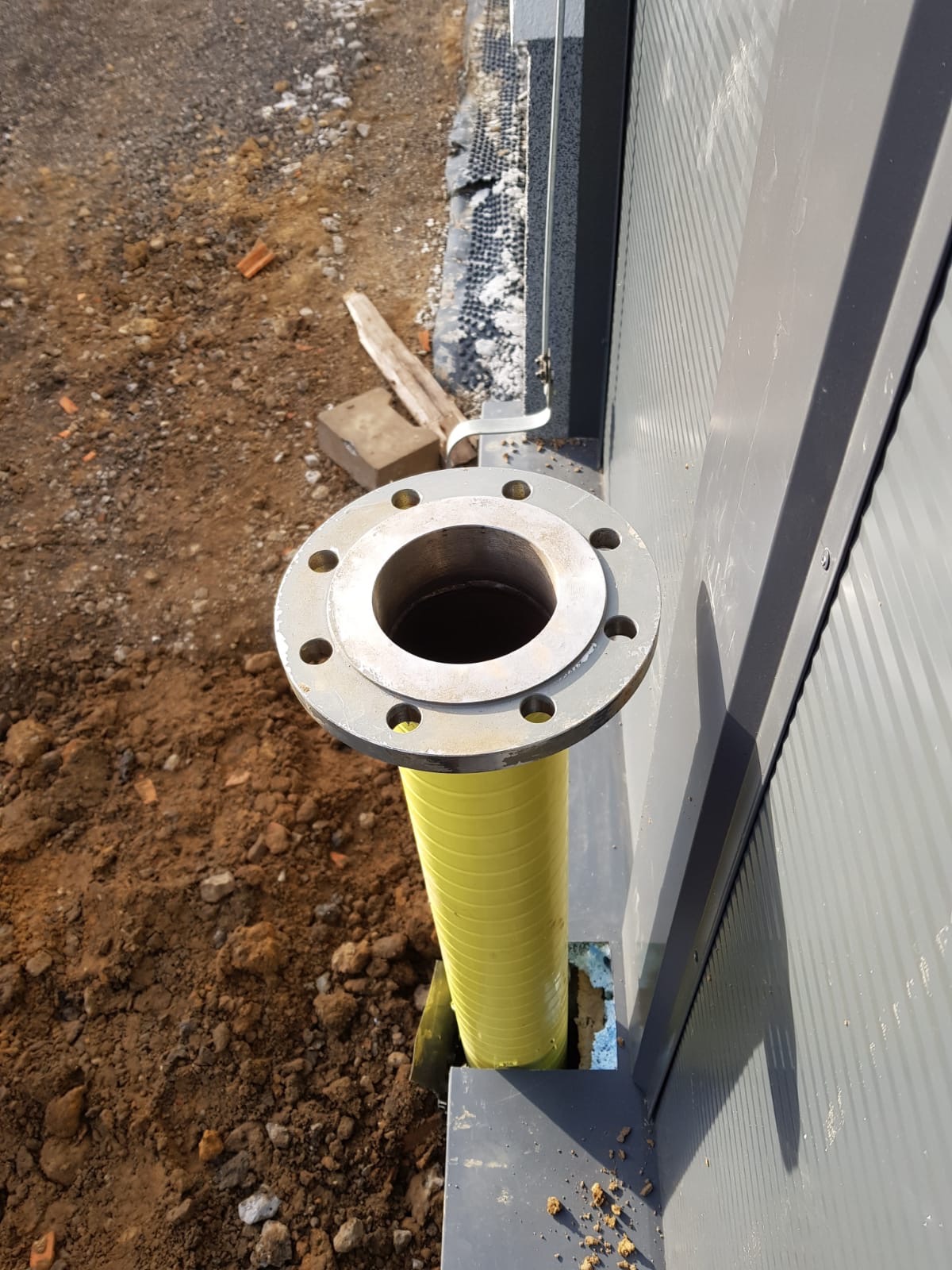 Żółta rura z kołnierzem wystająca z ziemi przy ścianie budynku. Widoczne elementy instalacji i ziemia.