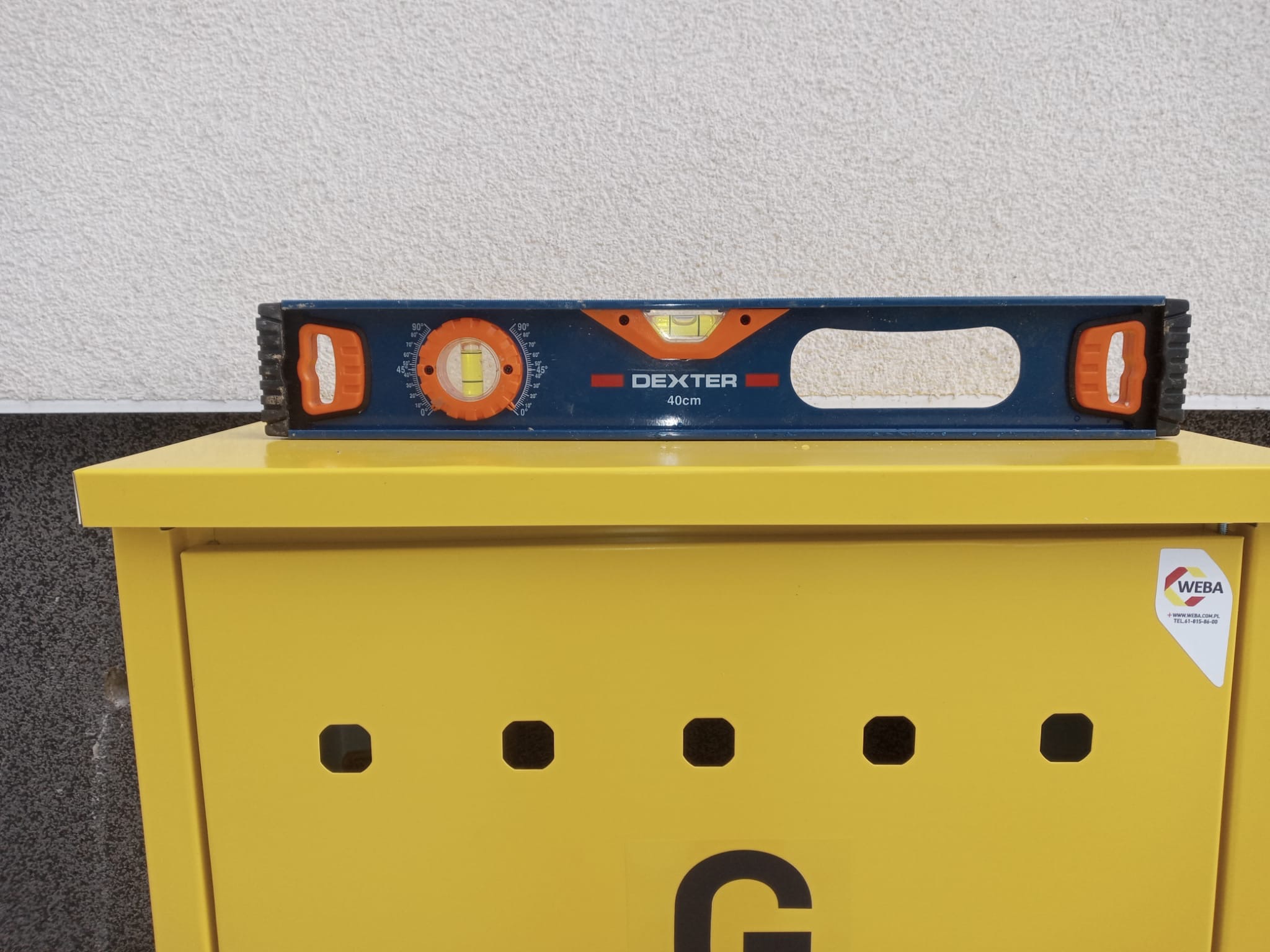 Niebieska poziomica Dexter 40cm leży na żółtej szafce z otworami wentylacyjnymi, na tle białej ściany o fakturze baranka. Widoczne logo WEBA.