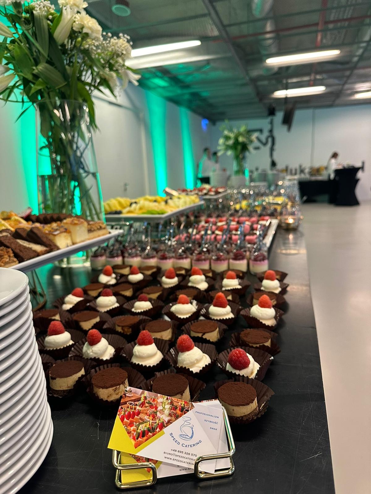 Stół z deserami: mini torciki z kremem i maliną, ciasta, owoce. W tle białe kwiaty w wazonach i zielone oświetlenie. Na pierwszym planie wizytówka firmy cateringowej.