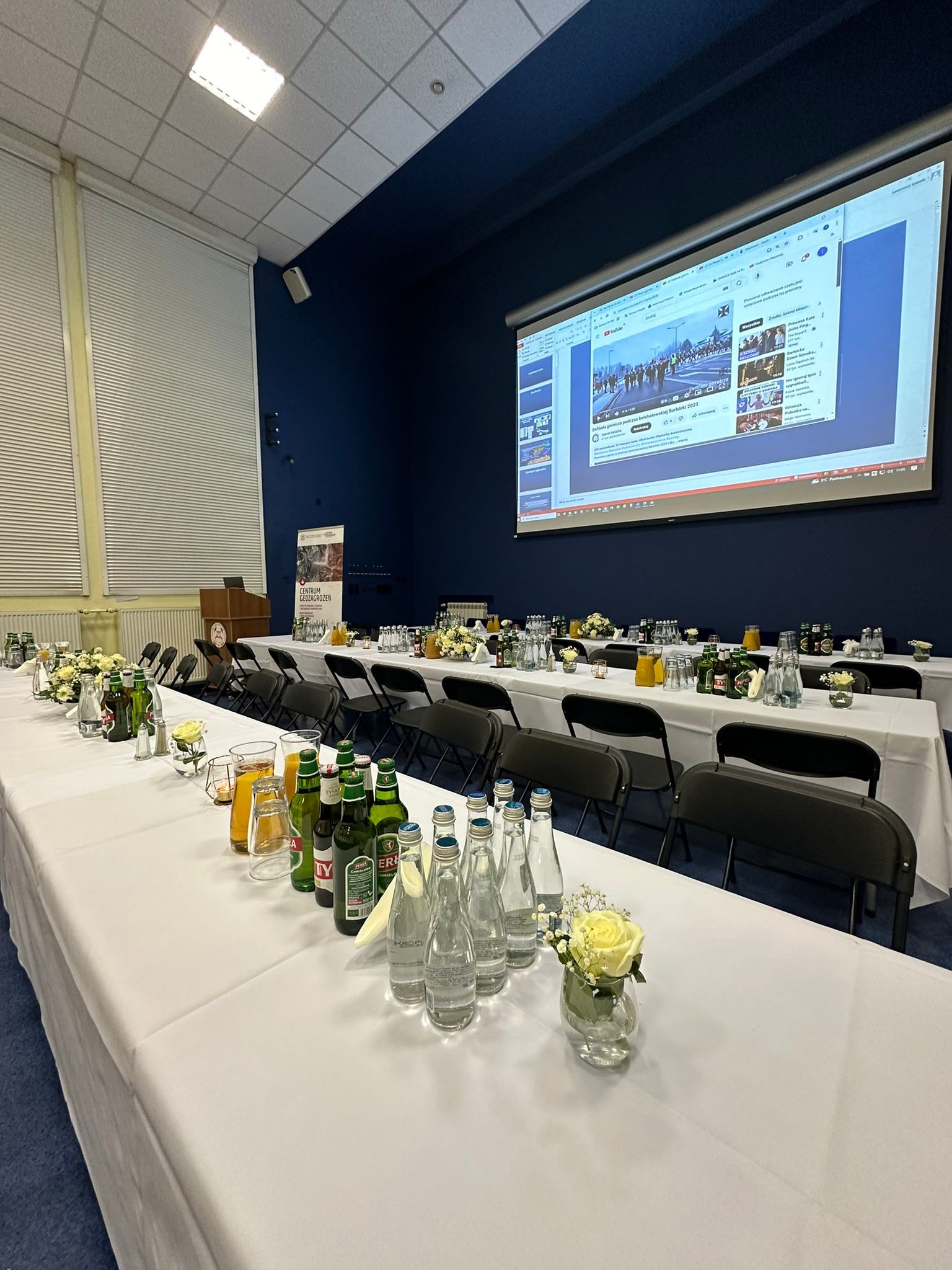 Długi stół nakryty białym obrusem, przygotowany na konferencję lub bankiet, z napojami, szklankami, dekoracjami z białych róż i krzesłami; w tle ekran projekcyjny.