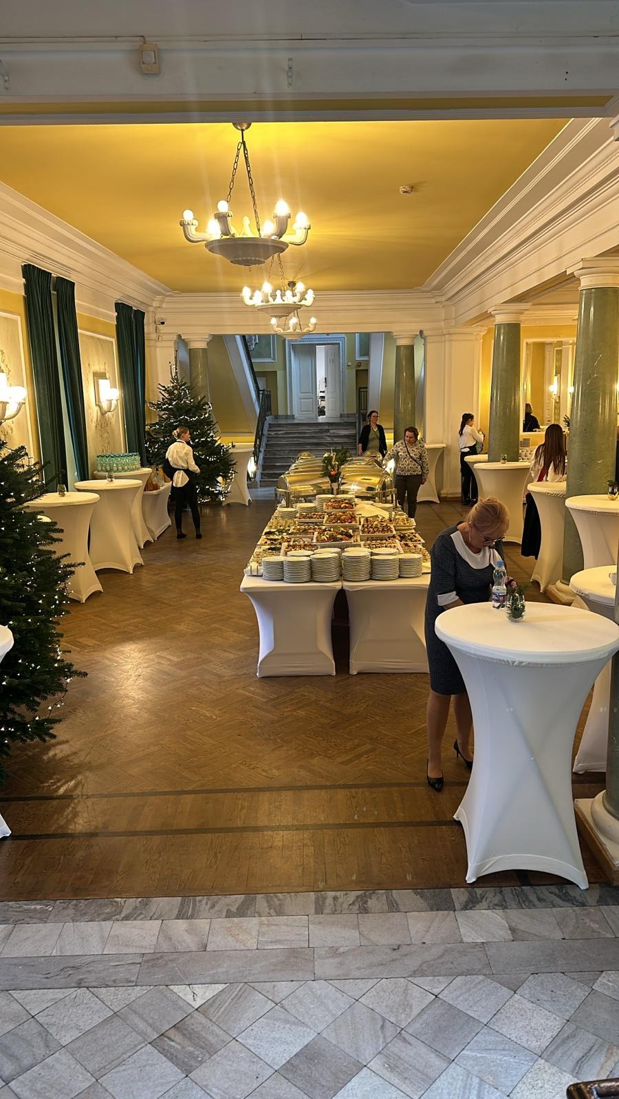 Sala bankietowa z długim stołem bufetowym pełnym potraw, białymi obrusami i dekoracjami świątecznymi, w tle widoczni goście.