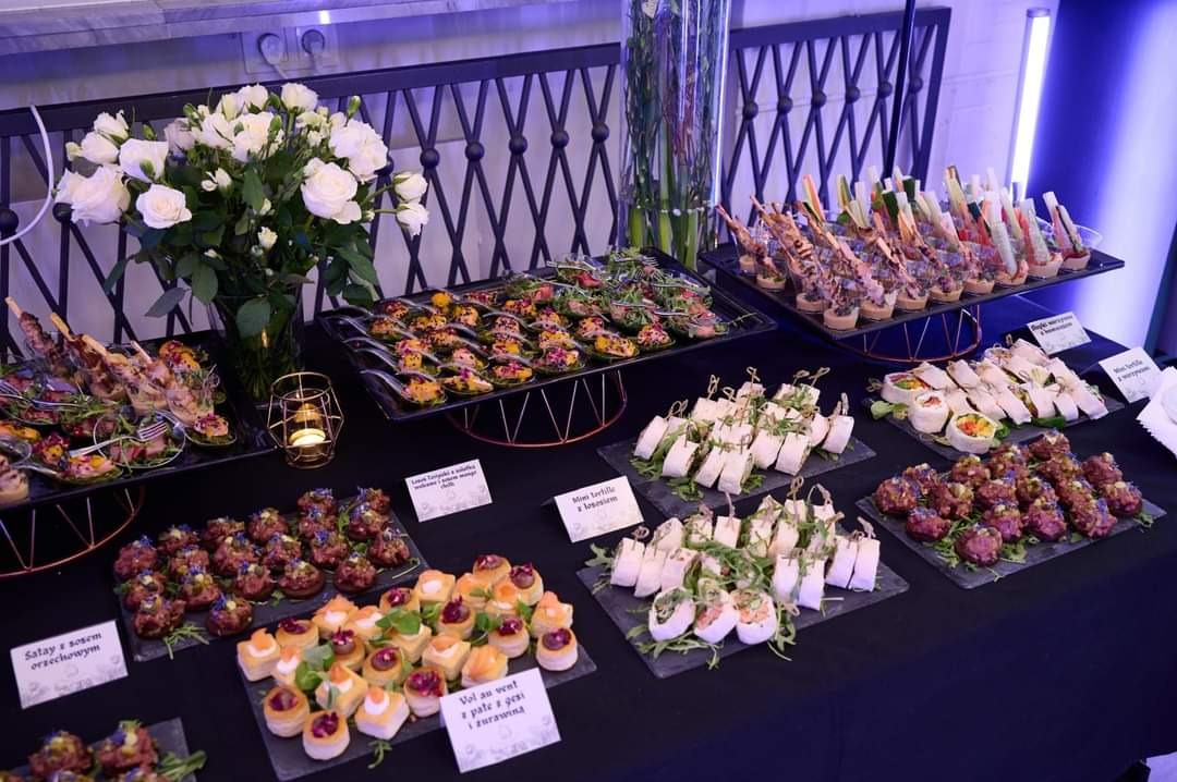 Stół z różnorodnymi przekąskami: koreczki, mini tortille, sałatki w łyżeczkach, vol-au-vent, dekorowany białymi różami i świecami, ustawiony na czarnym obrusie z metalowym ażurowym tłem.