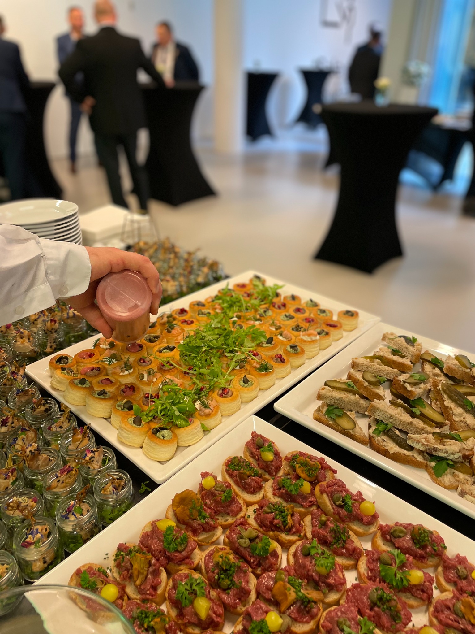 Różnorodne przekąski na białych prostokątnych tacach: tartinki z tatarem i kaparami, mini-kanapeczki z ogórkiem kiszonym, babeczki z sałatką i dekoracją z zieleniny, oraz małe słoiczki z kompozycjami.