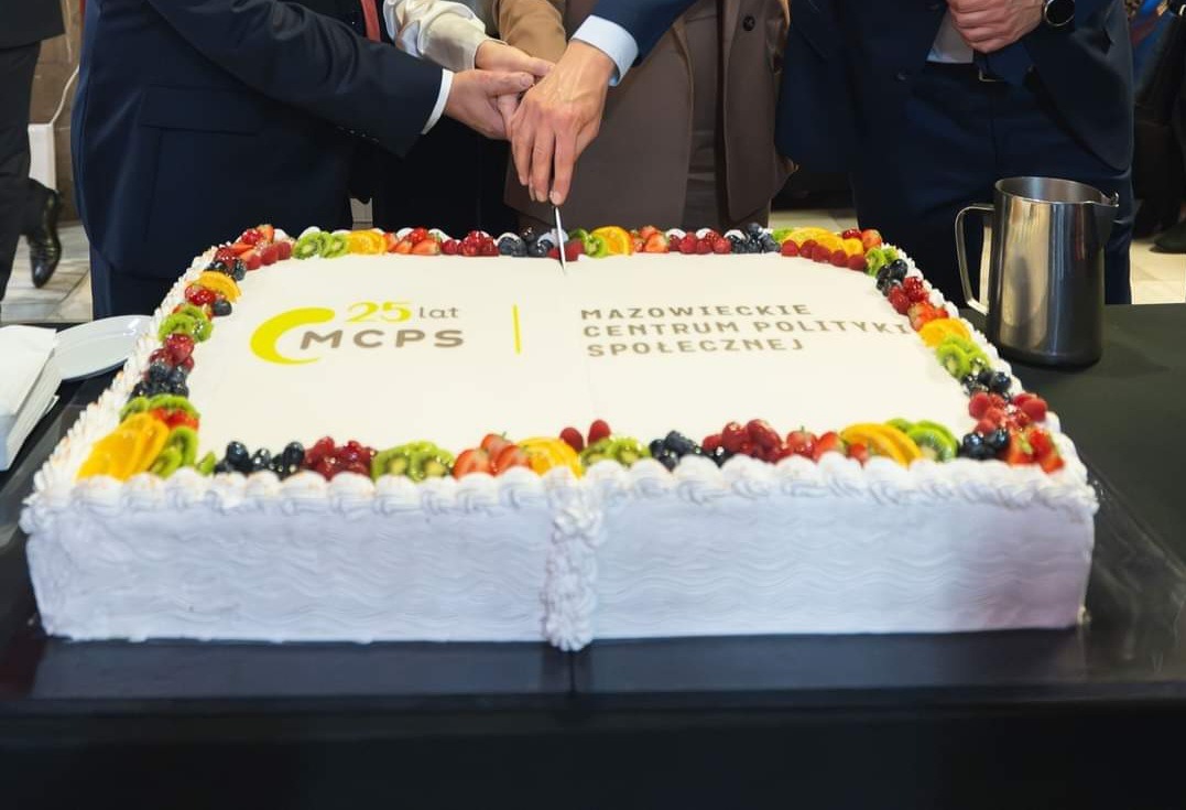 Krojenie dużego, kwadratowego tortu z napisem '25 lat MCPS Mazowieckie Centrum Polityki Społecznej', ozdobionego owocami, przez kilka osób w garniturach.
