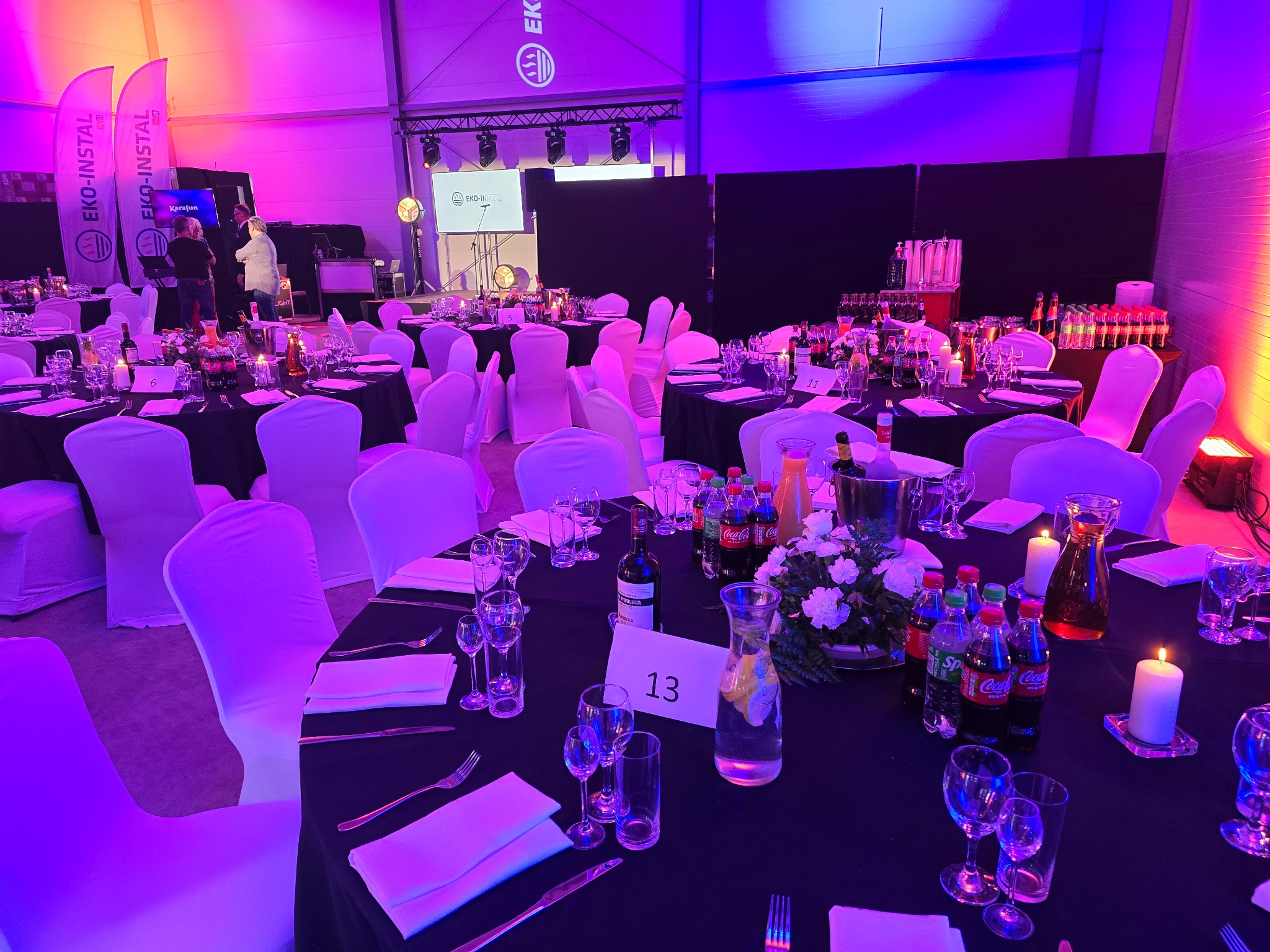 Uroczysta sala bankietowa z czarnymi obrusami, białymi pokrowcami na krzesłach, zastawionymi stołami z napojami, kwiatami i świecami, oświetlona różowo-fioletowym światłem, widoczny numer stołu 13.