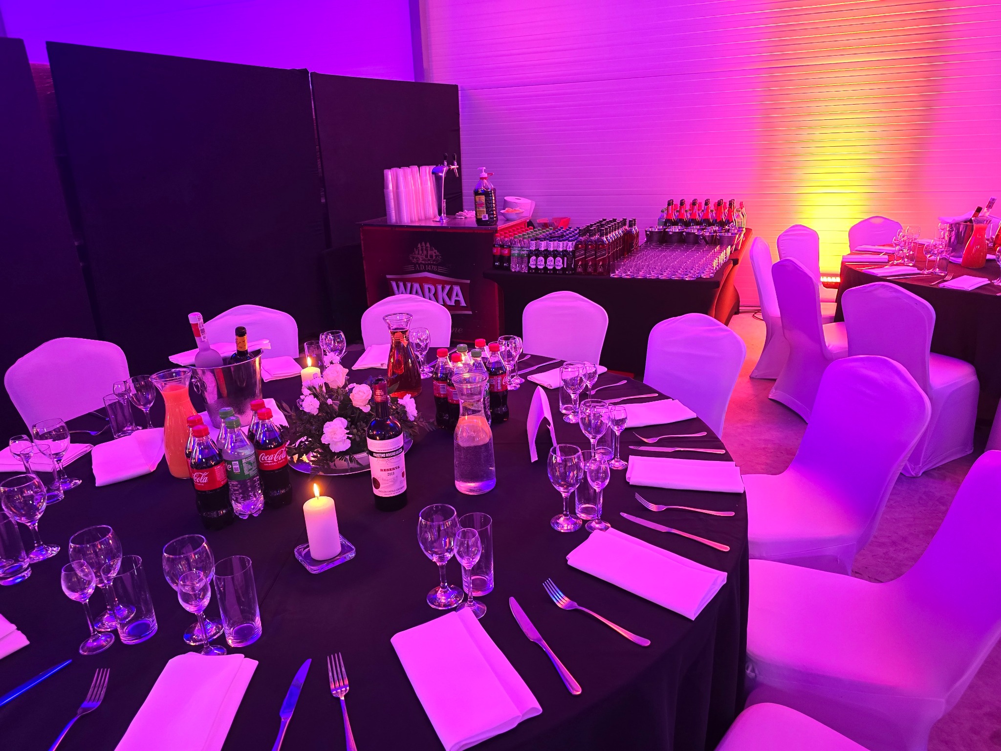Uroczysty stół z czarnym obrusem, nakryty zastawą, napojami i świecami, oświetlony fioletowo-różowym światłem, w tle bar z butelkami i szklankami.