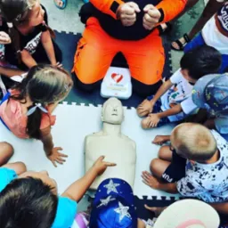 Grupa dzieci otacza manekina do resuscytacji na macie, instruktor w pomarańczowym kombinezonie pokazuje ćwiczenie, obok defibrylator.