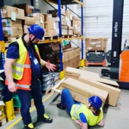 Instruktor demonstruje technikę uwalniania spod ciężaru podczas szkolenia w magazynie, pracownica w kasku i kamizelce leży pod kartonami.