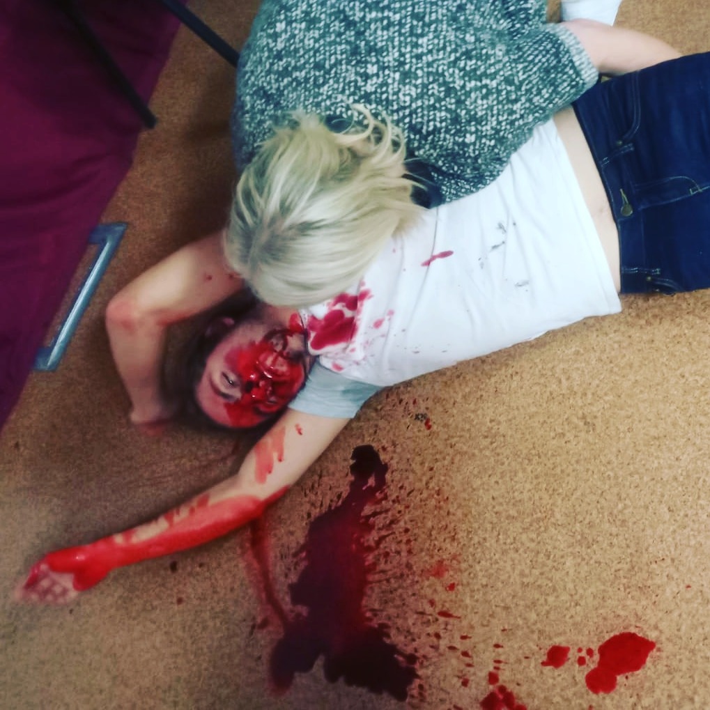 Symulacja udzielania pierwszej pomocy osobie z obrażeniami twarzy podczas szkolenia, widoczne sztuczne rany i krew, osoba udzielająca pomocy w swetrze klęczy obok poszkodowanego.