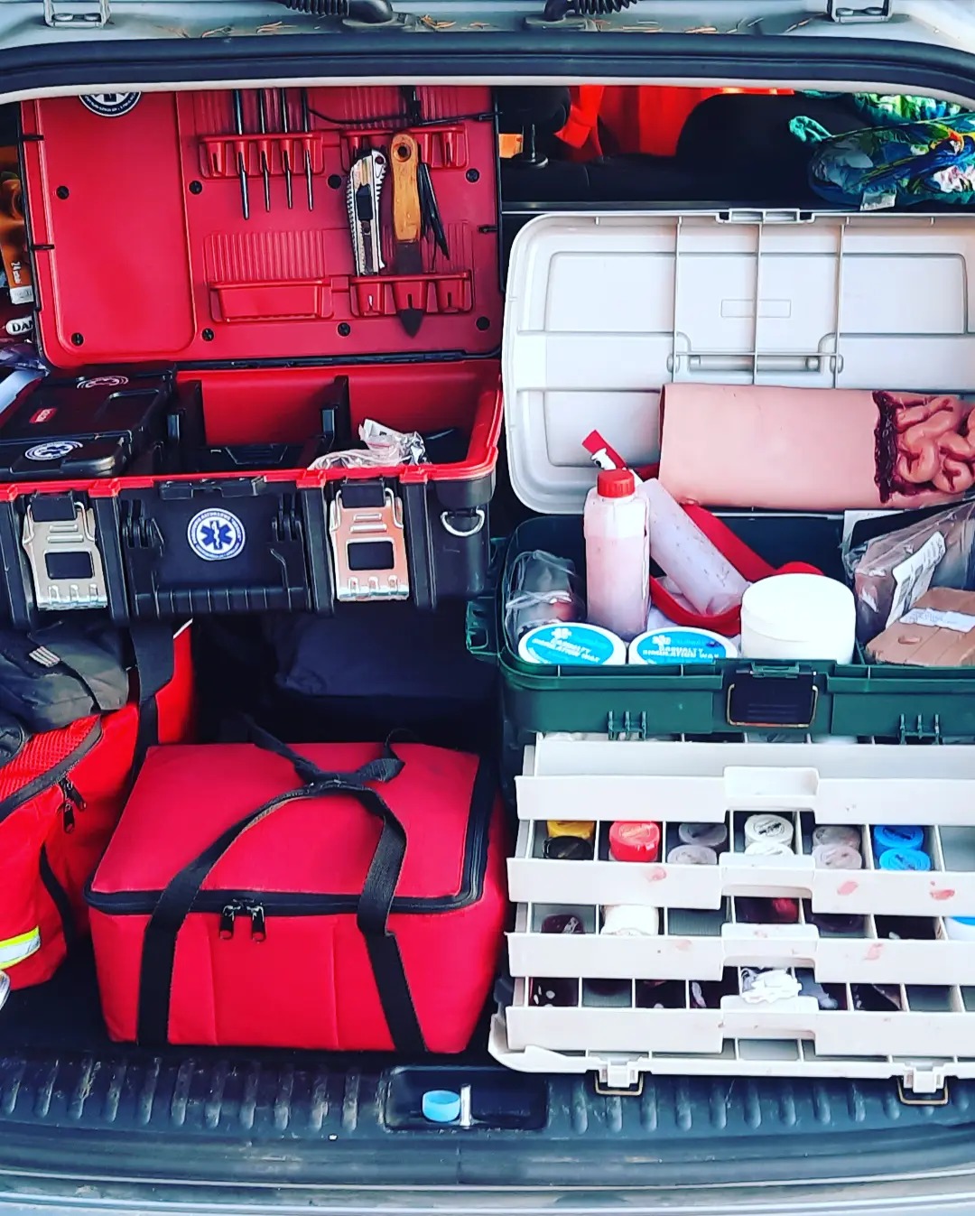 Otwarty bagażnik samochodu wypełniony sprzętem medycznym i szkoleniowym, w tym manekin z widocznymi wnętrznościami, apteczki, narzędzia i materiały opatrunkowe.