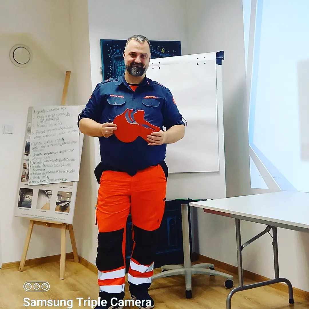 Instruktor w uniformie medycznym prezentuje model płuc podczas kursu pierwszej pomocy, w tle tablica z notatkami i projektor.