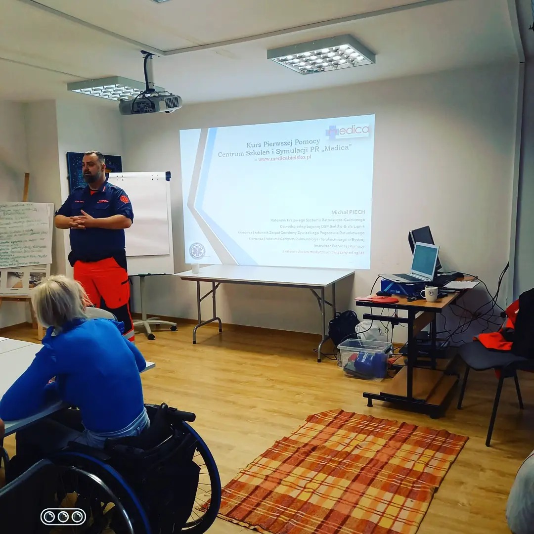 Instruktor w stroju ratowniczym prowadzi kurs pierwszej pomocy. Na ekranie projektora wyświetlona prezentacja z nazwą kursu i logiem Medica. Uczestniczka kursu na wózku inwalidzkim.