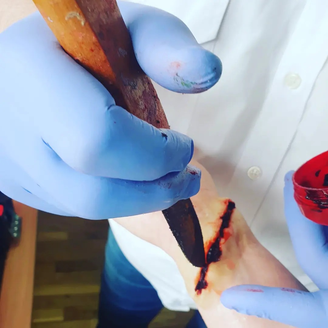 Nałożenie sztucznej rany na rękę przy użyciu drewnianej szpatułki i sztucznej krwi, osoba w niebieskich rękawiczkach i białej koszuli.