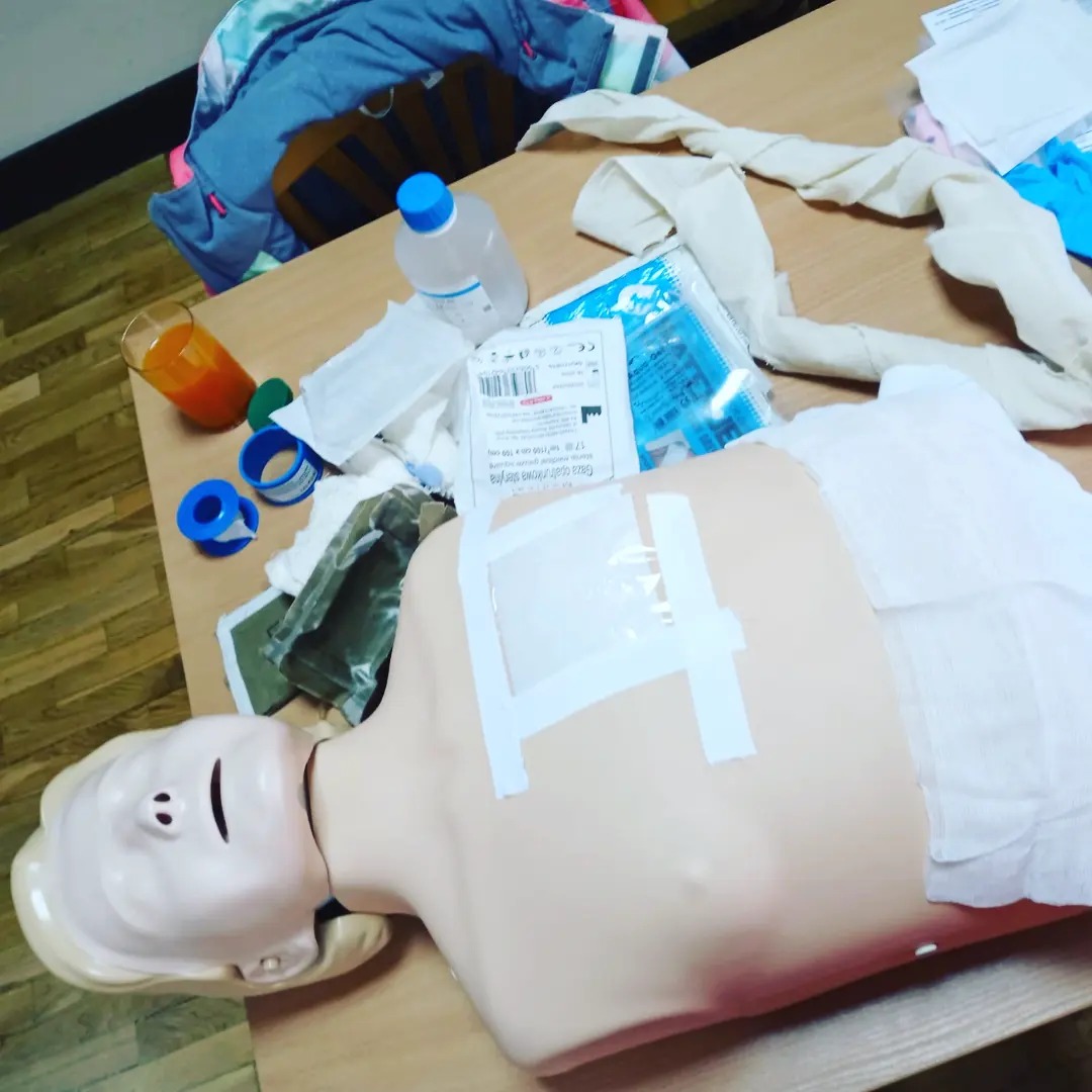 Manekin do nauki pierwszej pomocy z opatrunkiem na klatce piersiowej, leżący na stole obok materiałów opatrunkowych, płynu dezynfekującego i szklanki z sokiem.