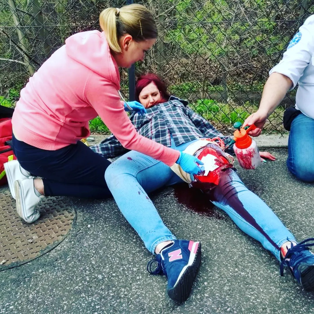 Symulacja udzielania pierwszej pomocy osobie z krwawiącą raną nogi podczas kursu, widoczni instruktorzy w rękawiczkach i osoba poszkodowana w jeansach i koszuli w kratę leżąca na chodniku.