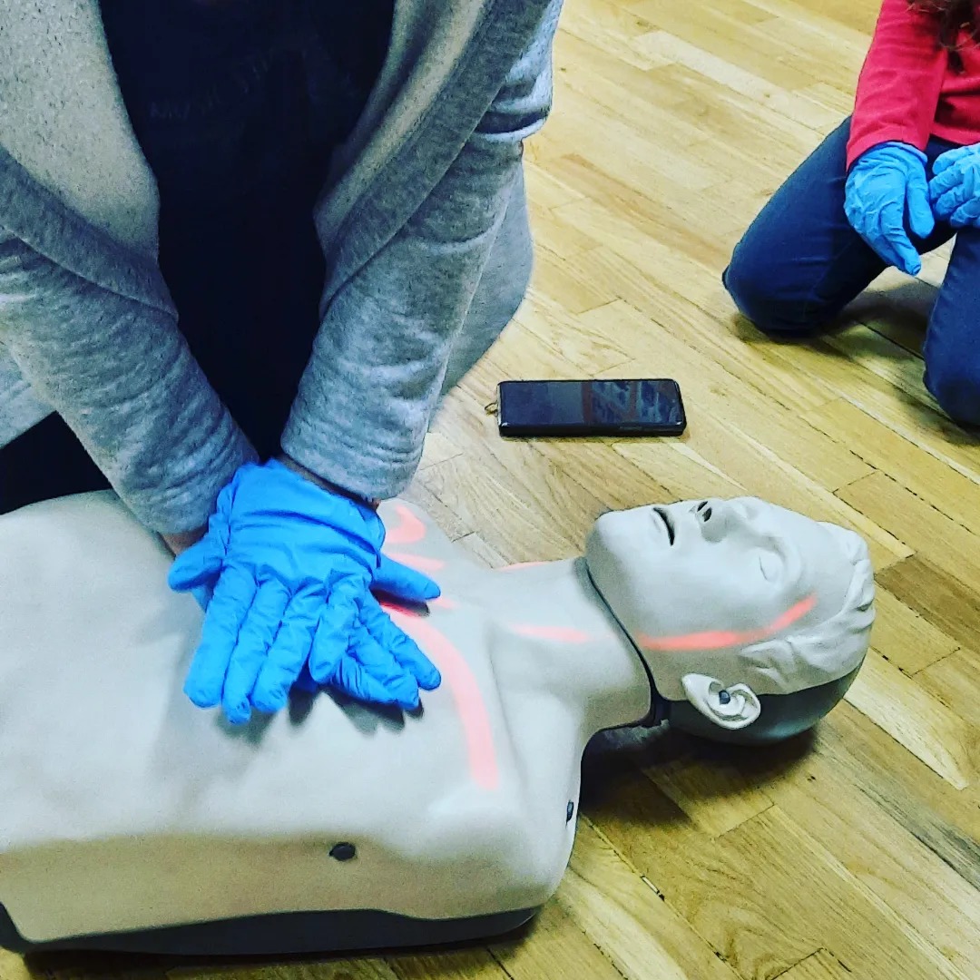 Manekin do resuscytacji krążeniowo-oddechowej z zaznaczonymi punktami ucisku, dłonie w niebieskich rękawiczkach wykonują masaż serca, obok leży telefon.