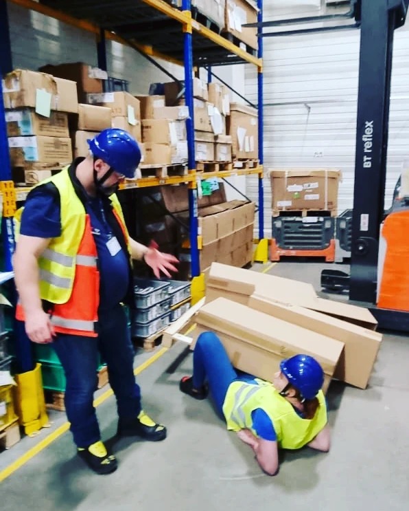 Instruktor demonstruje technikę uwalniania spod ciężaru podczas szkolenia w magazynie, pracownica w kasku i kamizelce leży pod kartonami.