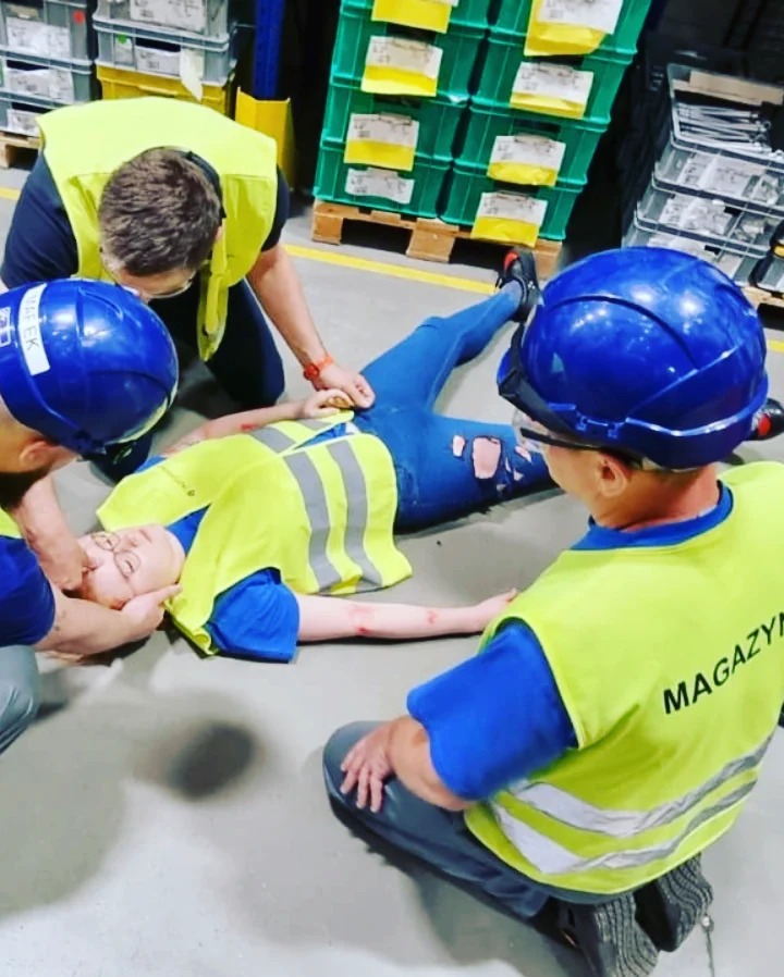 Symulacja udzielania pierwszej pomocy poszkodowanej z ranami na ręce i podartymi spodniami, w tle magazyn z zielonymi pojemnikami, uczestnicy w kaskach i kamizelkach odblaskowych.