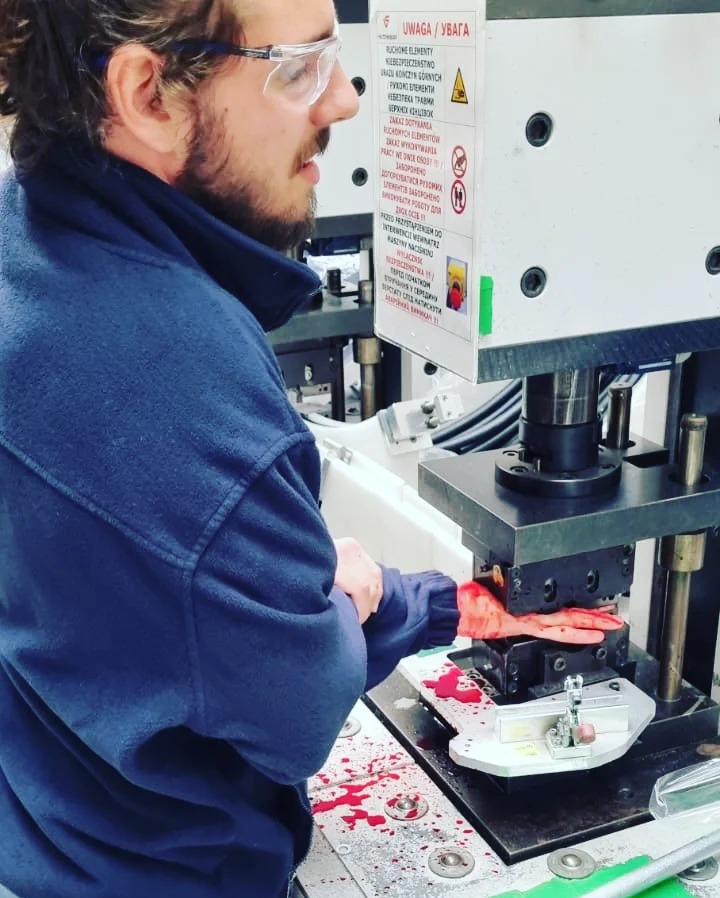 Mężczyzna w okularach ochronnych demonstruje działanie prasy z atrapą dłoni, na której widoczne są ślady imitujące krew, obok maszyny naklejka ostrzegawcza.