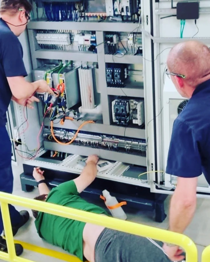 Trzech mężczyzn pracuje przy dużej szafie sterowniczej: jeden leży na podłodze i coś podłącza, drugi sprawdza połączenia, a trzeci nadzoruje pracę.