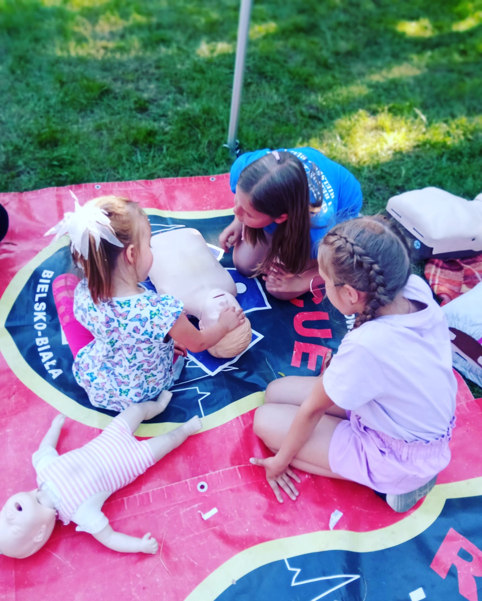 Trzy dziewczynki ćwiczą resuscytację krążeniowo-oddechową na manekinie podczas kursu pierwszej pomocy na trawie, obok leży lalka.