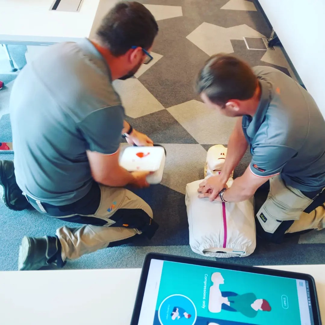 Dwaj mężczyźni w szarych koszulkach demonstrujący resuscytację krążeniowo-oddechową na manekinie, z otwartym pojemnikiem z akcesoriami i tabletem wyświetlającym instrukcje na pierwszym planie,...