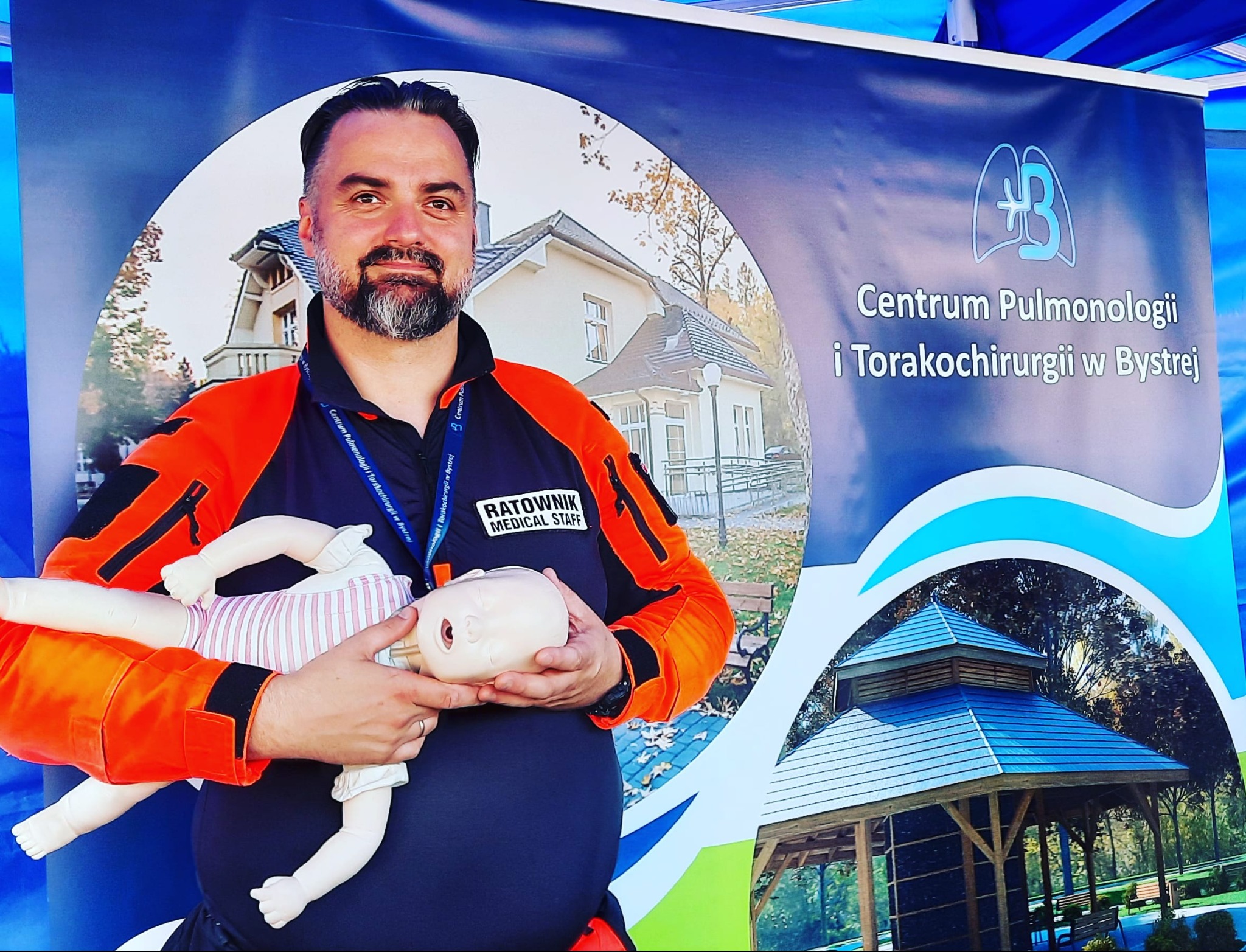 Ratownik medyczny w pomarańczowo-granatowym uniformie prezentuje manekin niemowlęcia na tle baneru Centrum Pulmonologii i Torakochirurgii w Bystrej.