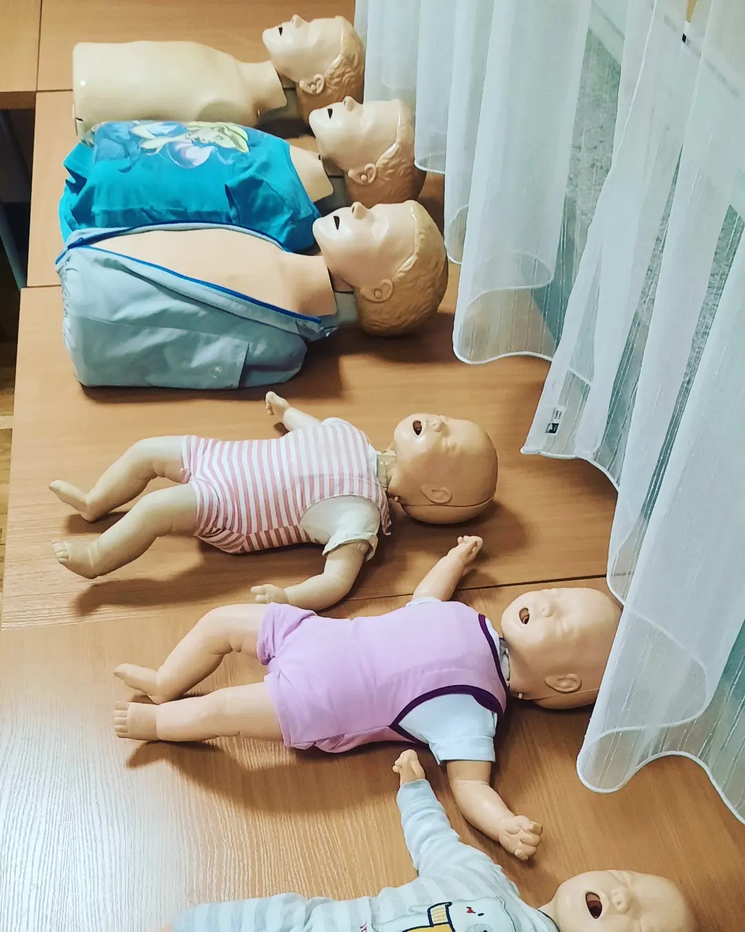 Manekiny do nauki resuscytacji ułożone na drewnianym stole, widoczne manekiny dorosłych i dzieci, tło stanowią białe, prześwitujące zasłony.