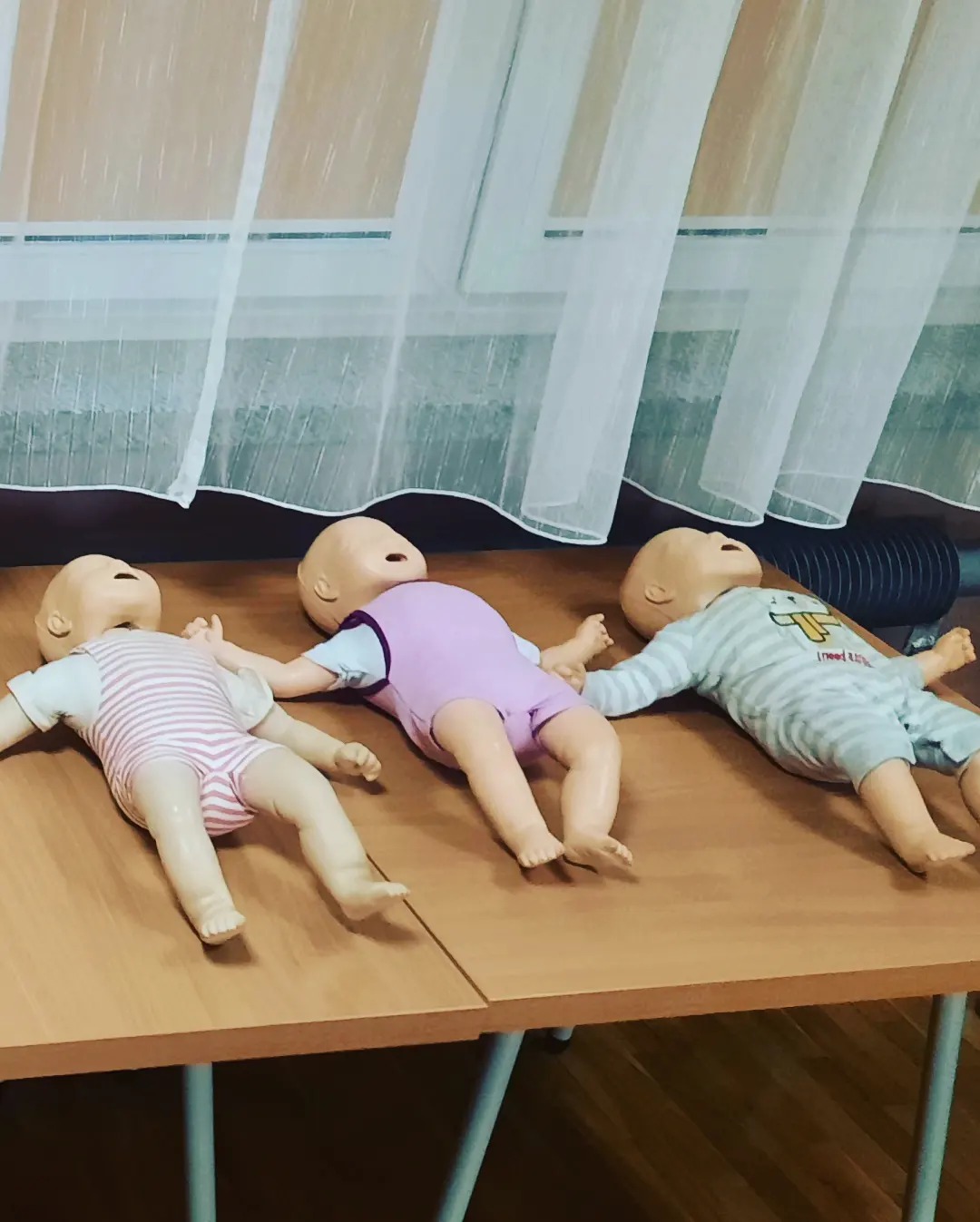 Trzy manekiny niemowlęce ułożone na stole, przygotowane do ćwiczeń resuscytacji, na tle okna z białą firanką.