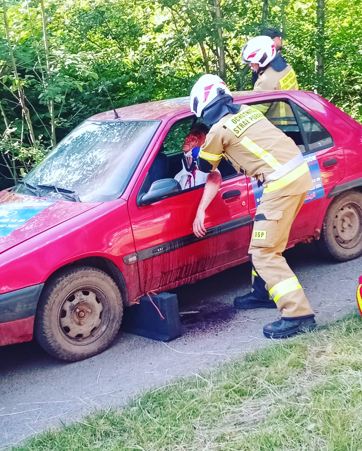 Symulacja akcji ratunkowej: strażacy OSP udzielają pomocy poszkodowanemu w czerwonym samochodzie osobowym po wypadku, widoczne ślady krwi.