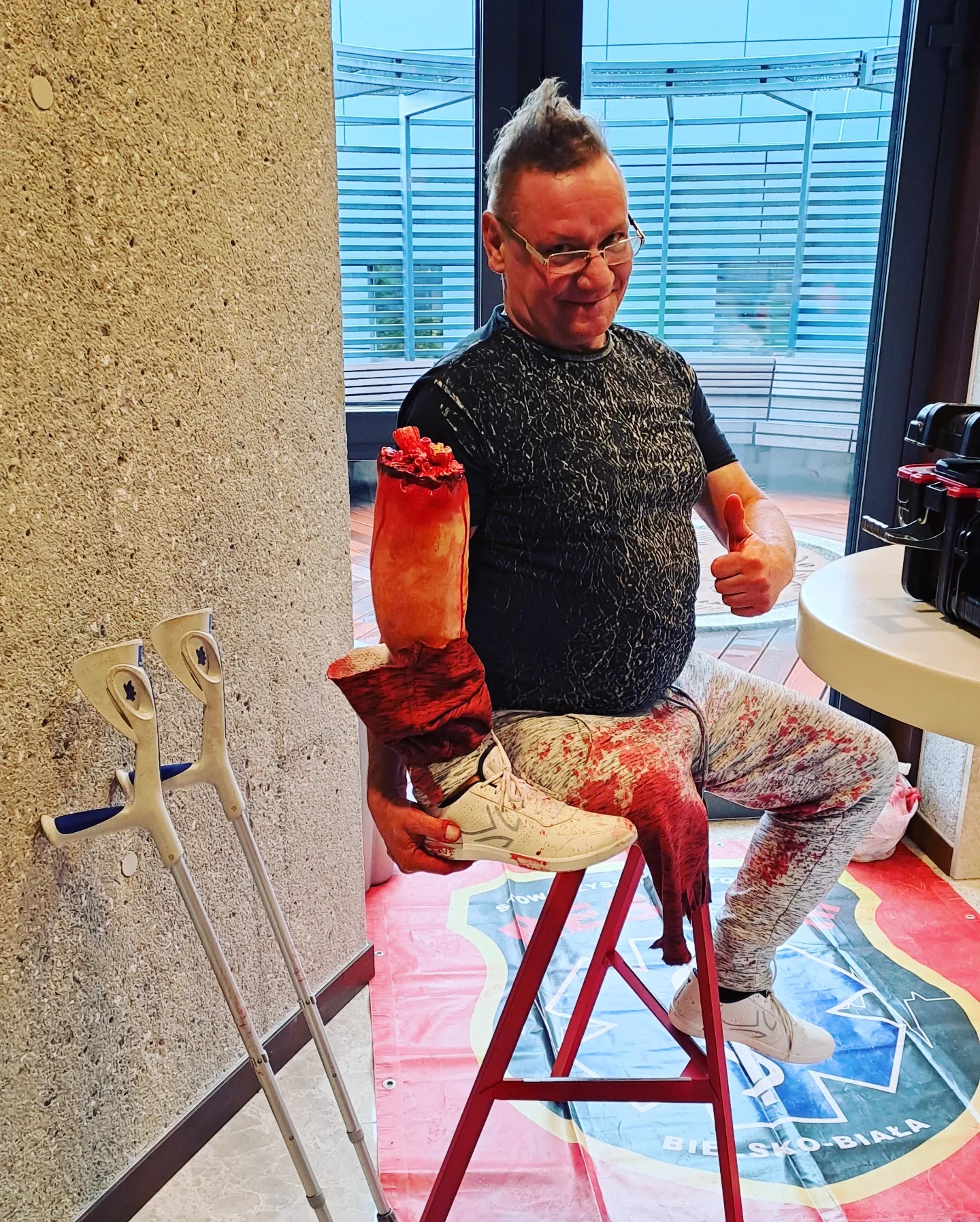 Mężczyzna z irokezem, w okularach, siedzi na czerwonym stołku z odciętą atrapą ręki i nogi pokrytą sztuczną krwią, obok kule ortopedyczne, kciuk w górę.