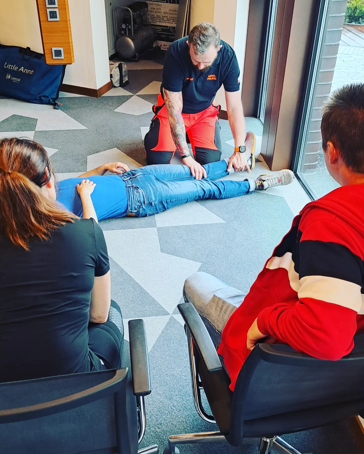 Instruktor w czerwonych spodniach i tatuażem na ramieniu demonstruje na leżącej osobie, jak prawidłowo badać puls na stopie, podczas gdy dwie osoby siedzą na krzesłach i obserwują.