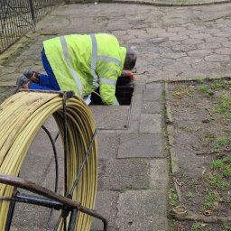 Braingeneration - Pracownik w odblaskowej kurtce pochylony nad studzienką kanalizacyjną na chodniku, obok zwój żółtego kabla na metalowym stojaku, w tle ulica z zaparkowanymi samochodami.