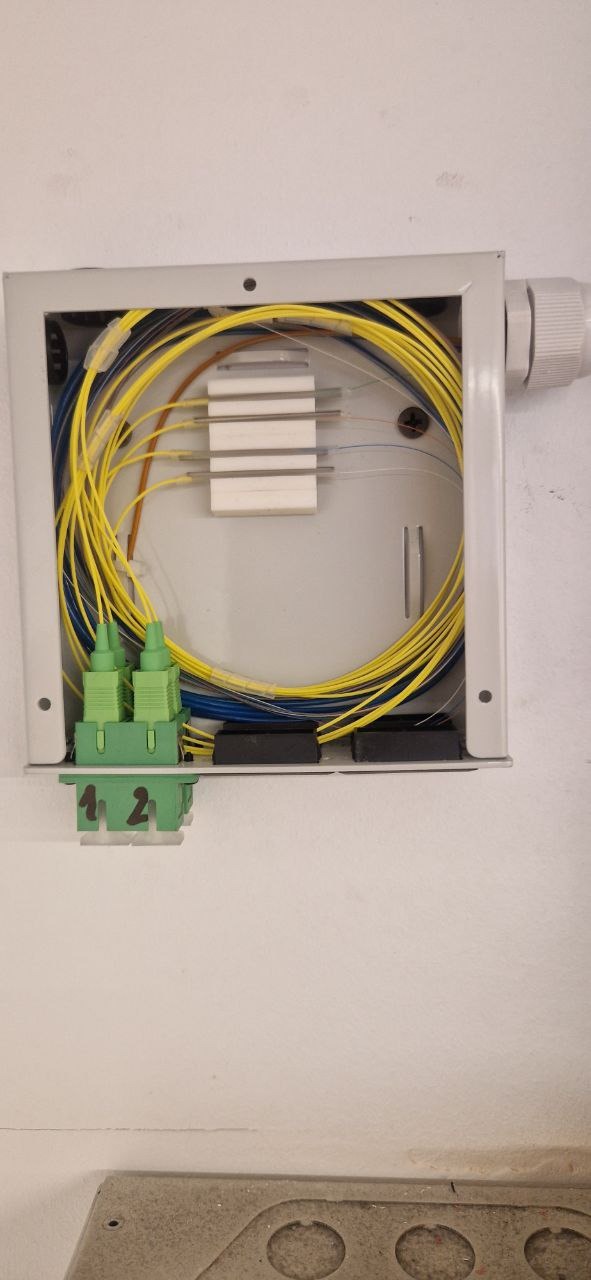 Otwarta puszka instalacyjna z żółtymi i niebieskimi kablami światłowodowymi, widoczne złącza i adaptery światłowodowe wewnątrz, puszka zamontowana na ścianie.