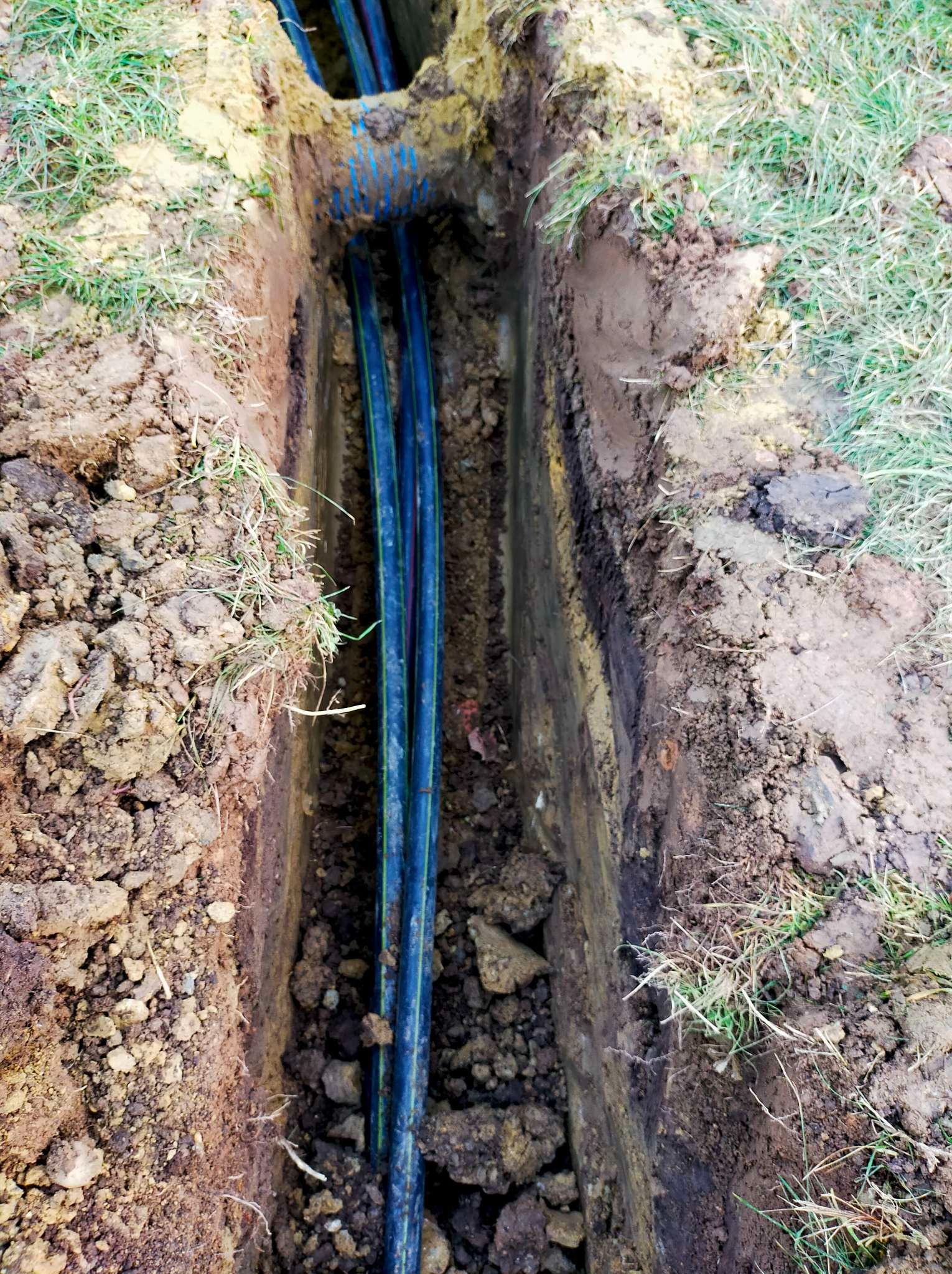 Wykop w ziemi z ułożonymi przewodami w niebieskiej osłonie karbowanej, widoczne warstwy gleby i trawa na powierzchni.