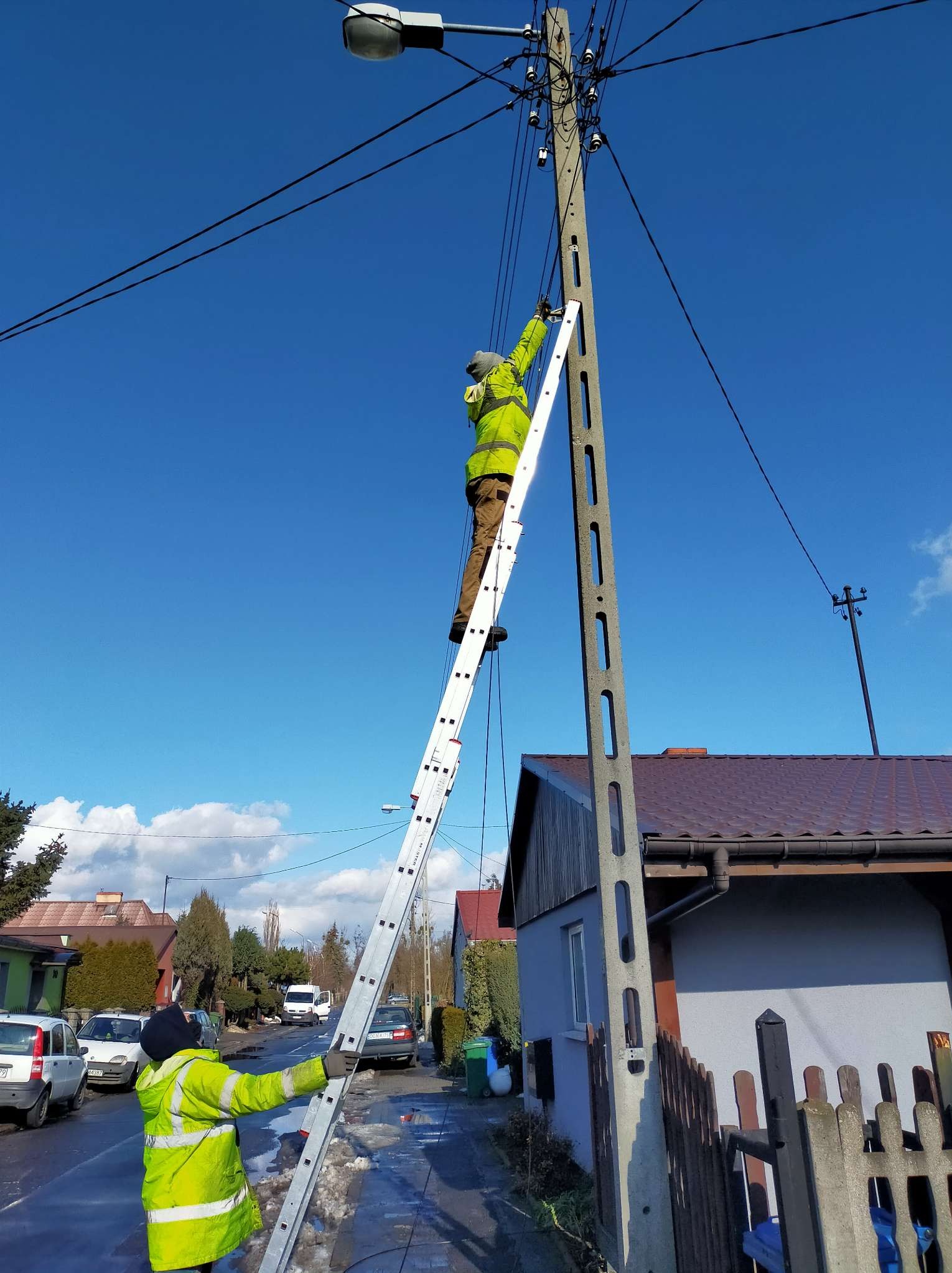 Dwóch pracowników w odblaskowych kurtkach pracuje przy słupie energetycznym na drabinie w słoneczny dzień, jeden asekuruje drabinę z dołu, drugi wykonuje prace na wysokości.