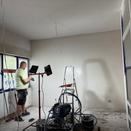 StagBud Sp z.o.o. - Prace przygotowawcze przed malowaniem w Szczecinie: mężczyzna szpachluje ścianę, widoczny agregat malarski, okna zabezpieczone folią.