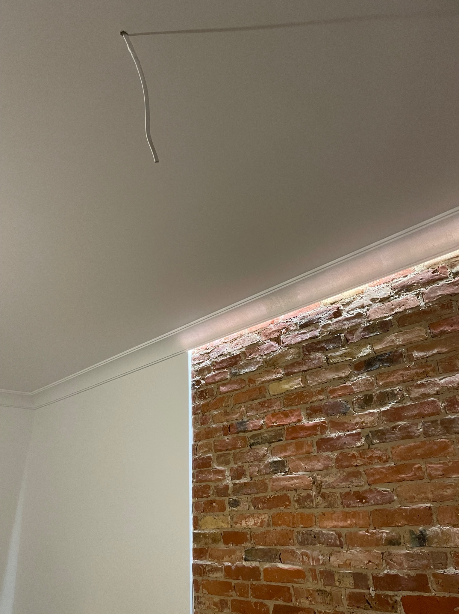 Fragment wnętrza z surową ceglaną ścianą, oświetleniem LED w listwie przysufitowej i świeżo pomalowaną ścianą, widoczny wystający kabel ze ściany.