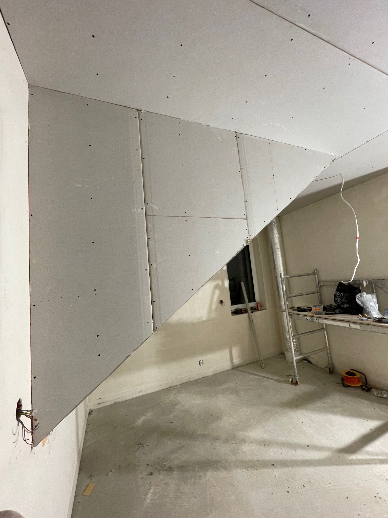 Montaż płyt gipsowo-kartonowych na skośnym poddaszu, widoczne łączenia płyt, szpachlowane otwory po wkrętach, drabina malarska w tle, surowa podłoga.