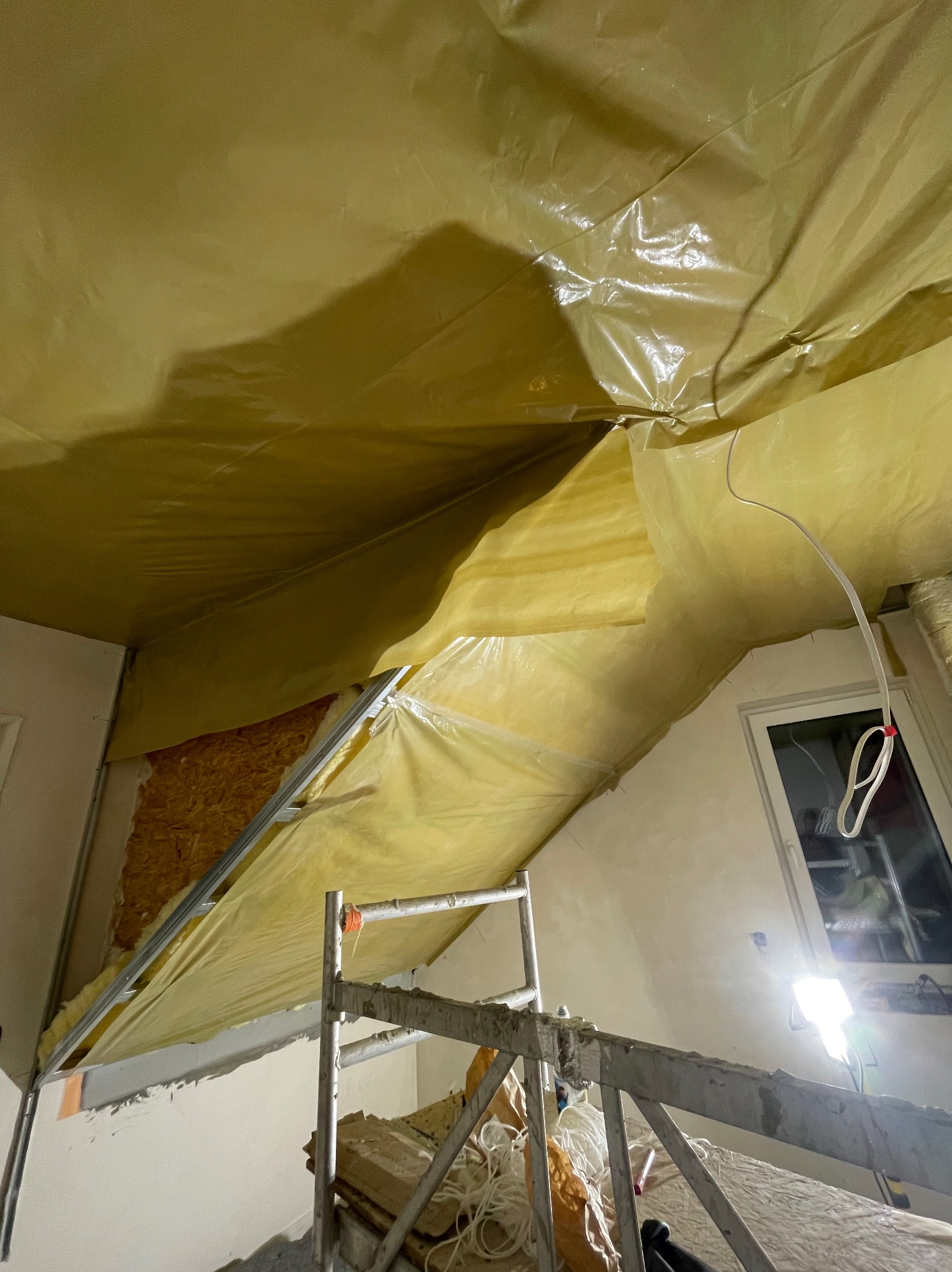 Przygotowanie poddasza do prac wykończeniowych: folia ochronna na suficie, widoczna drabina i oświetlenie robocze, fragment ściany z izolacją.