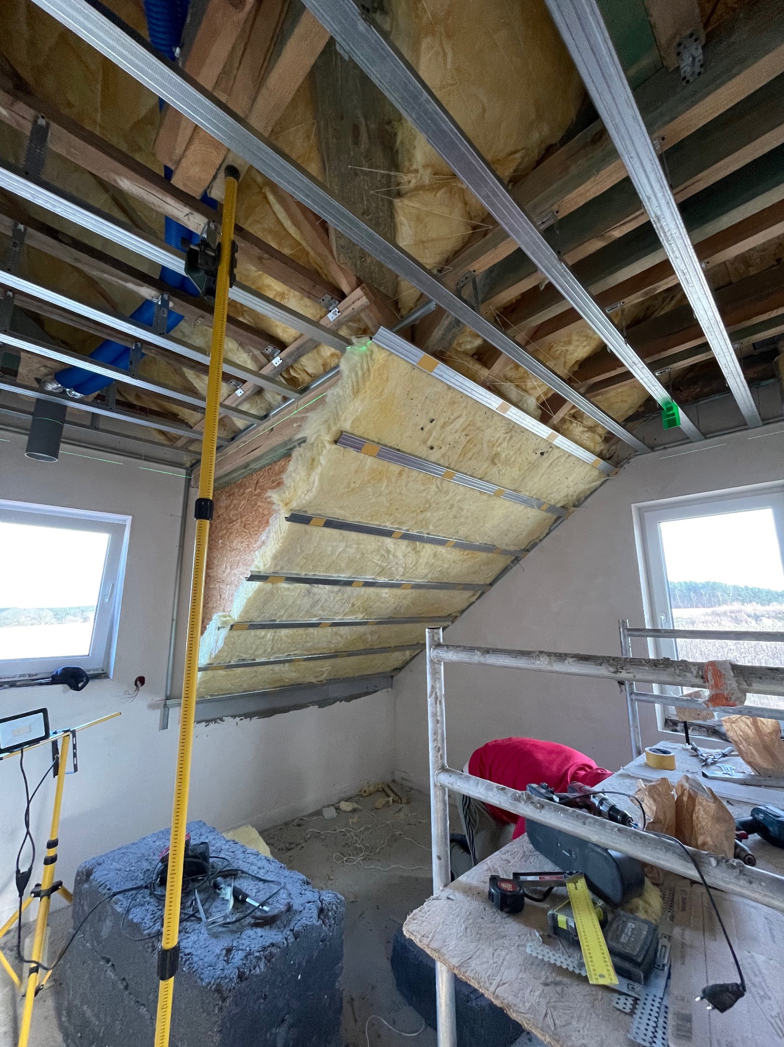 Ocieplenie poddasza wełną mineralną, widoczne metalowe profile, rusztowanie i narzędzia pracy, w tle okno z widokiem na drzewa.
