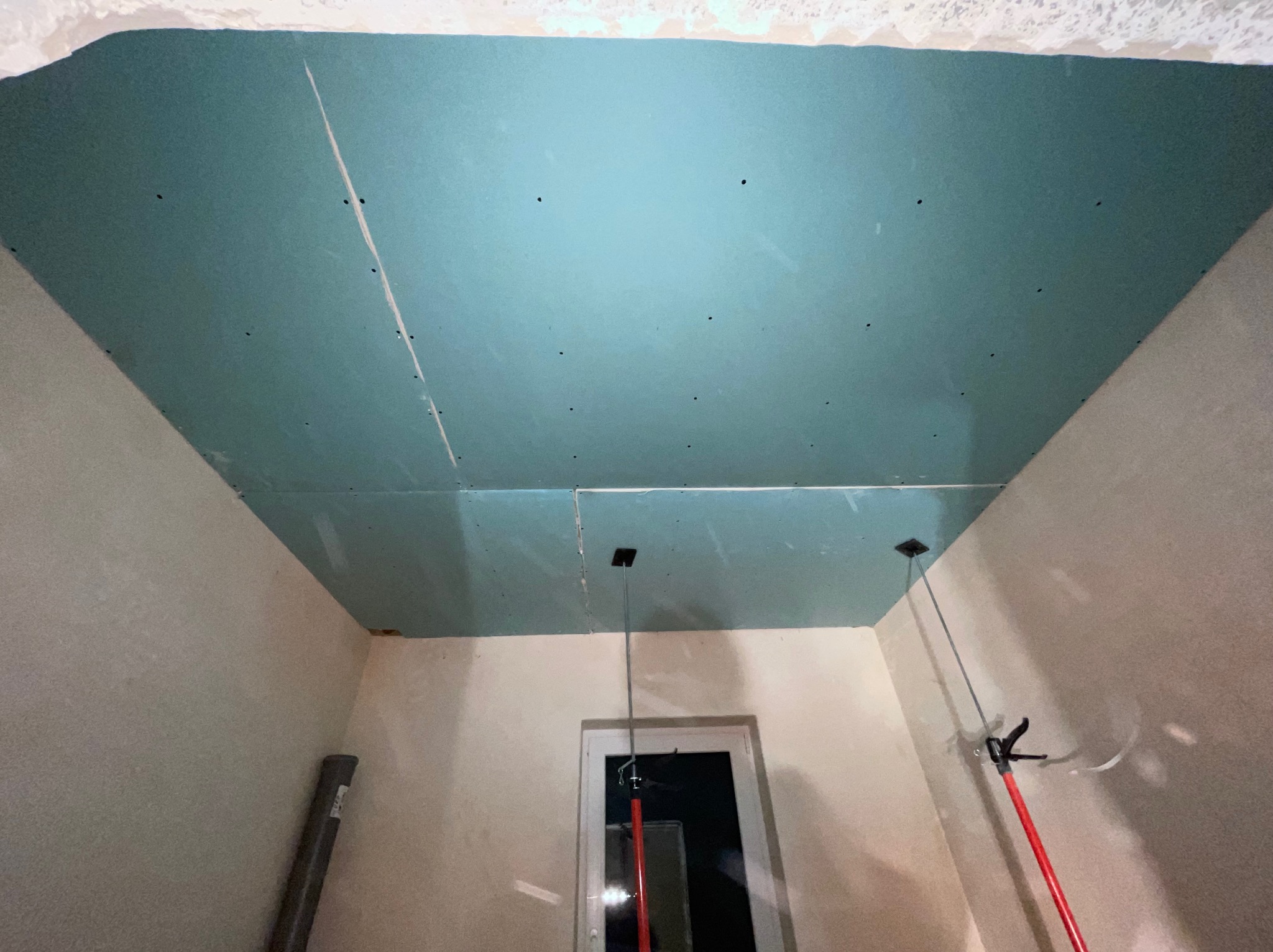 Montaż płyt gipsowo-kartonowych na suficie, widoczne łączenia i otwory na oświetlenie, wsporniki podtrzymujące konstrukcję, surowe ściany w tle.