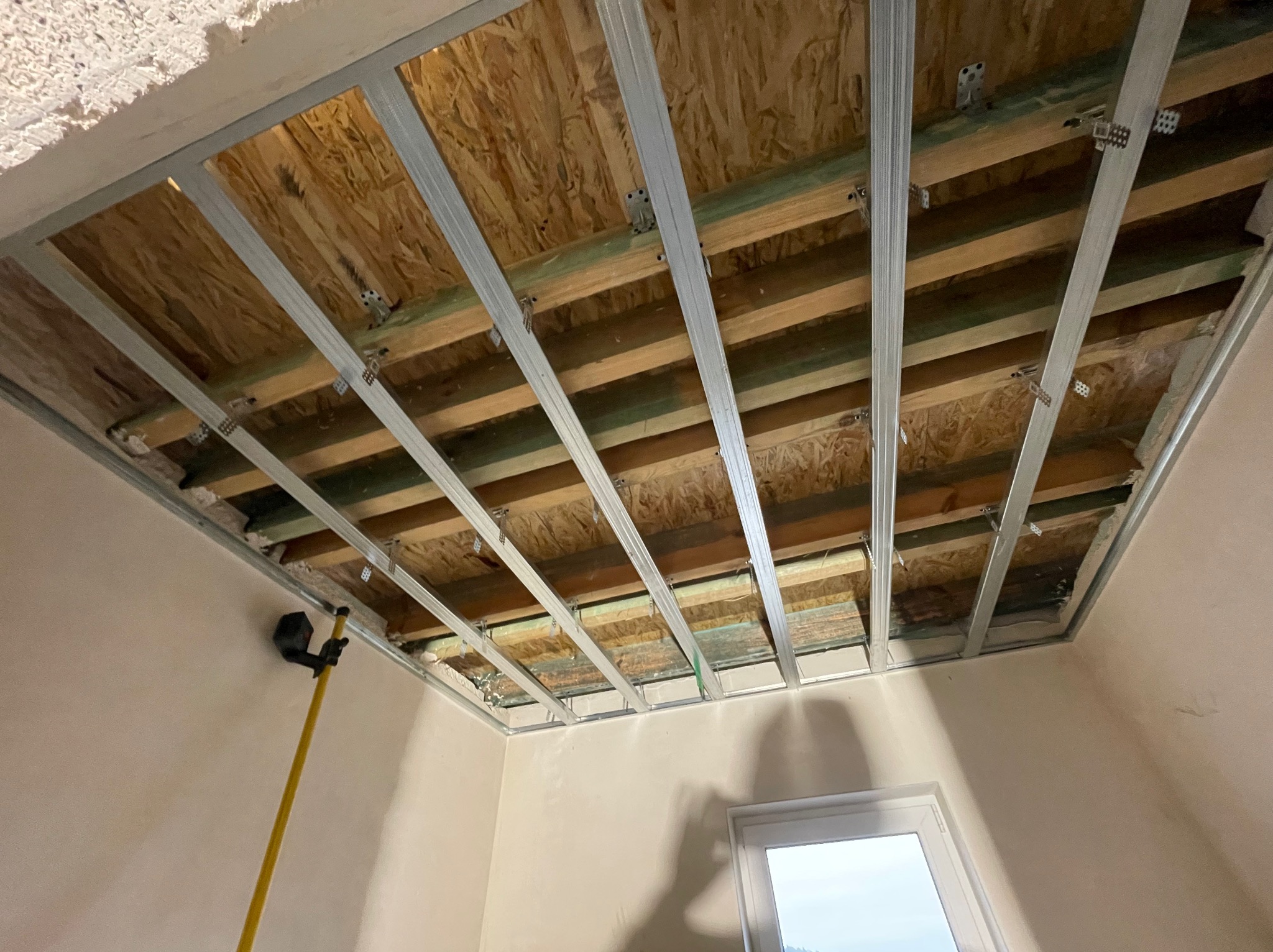 Metalowa konstrukcja sufitu podwieszanego z widocznymi elementami drewnianymi i płytami OSB, zamocowana do sufitu w pomieszczeniu z oknem i narzędziem do malowania ścian opartym o ścianę.