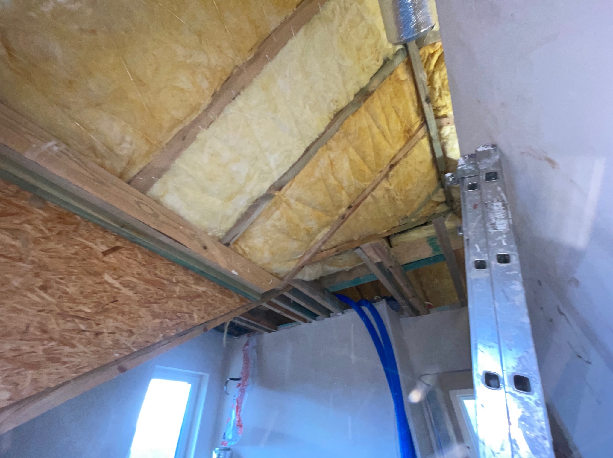 Poddasze w trakcie adaptacji: widoczne ocieplenie wełną mineralną między krokwiami, fragment płyty OSB, rury instalacji, oraz drabina oparta o ścianę.